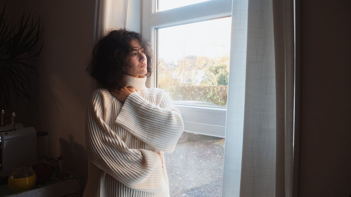 Wie versicherst du dich gegen Unwetterschäden? Frau mit weissem Pullover steht vor einem Fenster und guckt nachdenklich nach draussen.